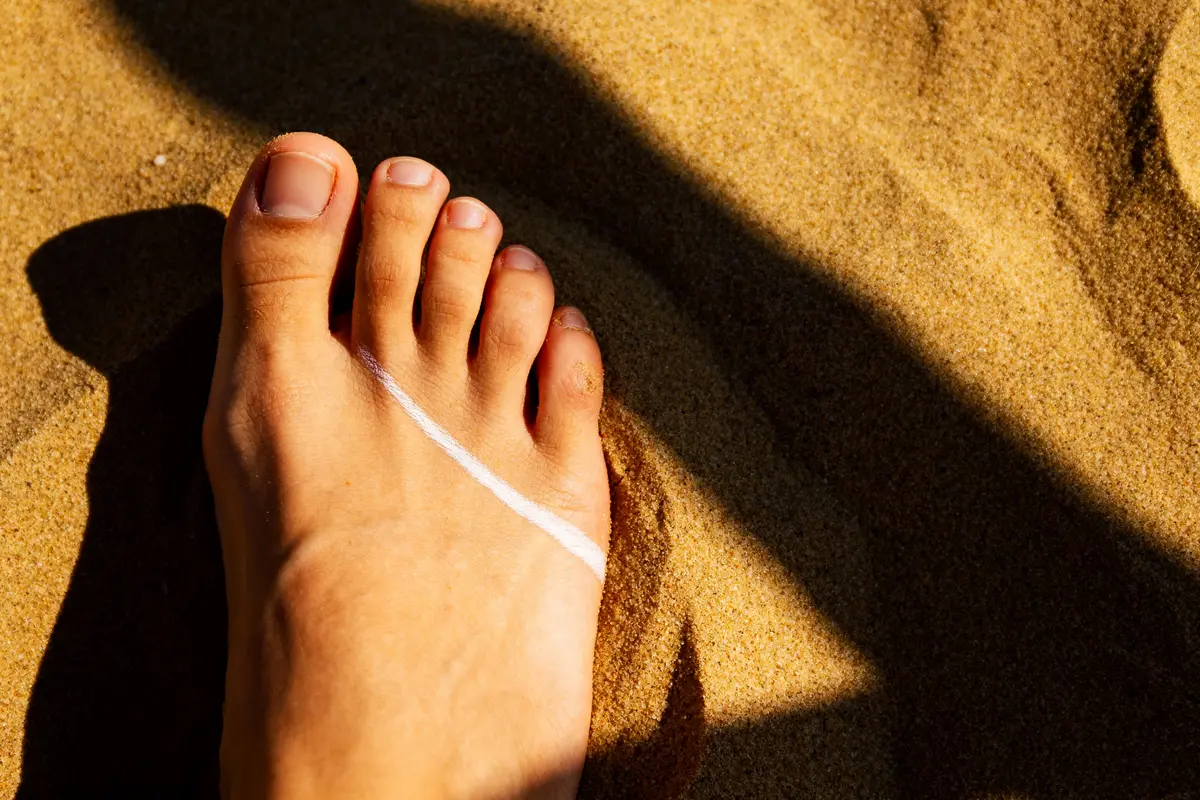 Pied nu sur sable, ligne de bronzage visible sous lumière ensoleillée.