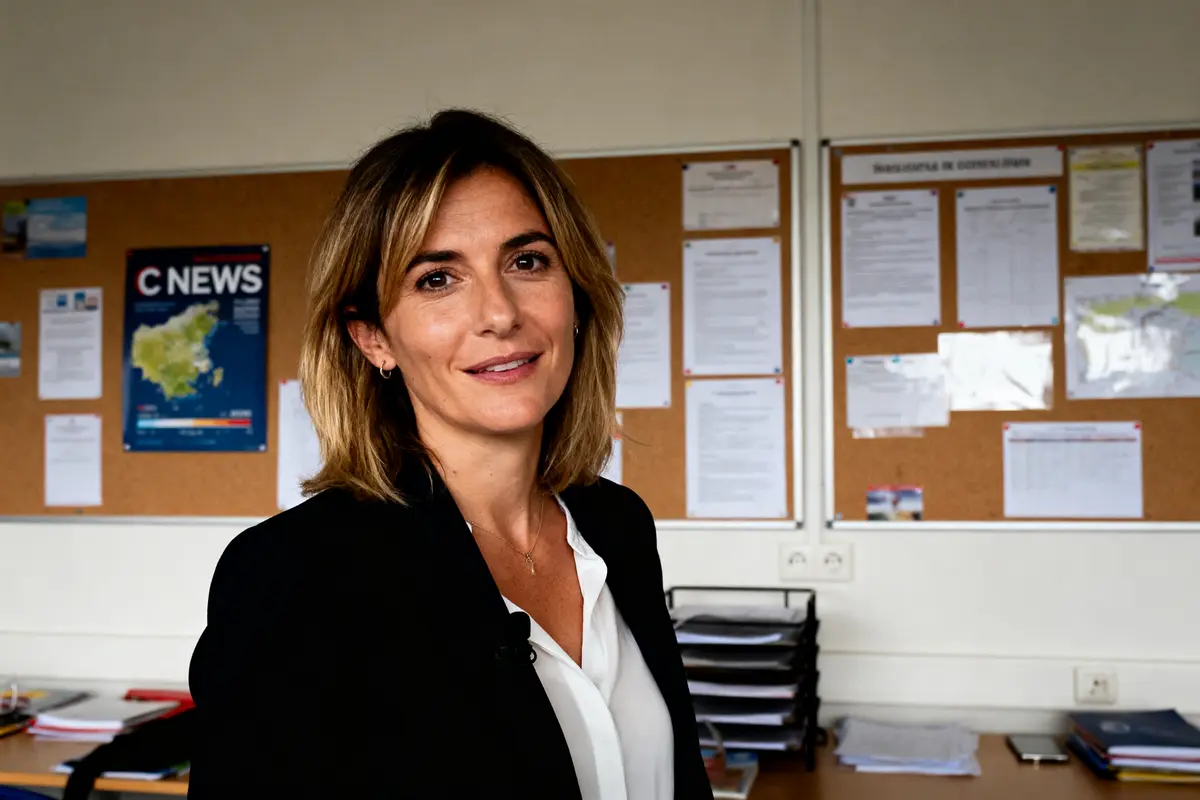 Femme souriante en veste noire devant un panneau d'affichage, documents et magazine "C News" visible.