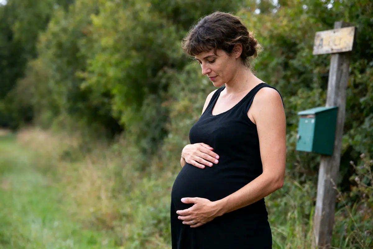 Femme enceinte en robe noire, posant dans un jardin, caressant son ventre, avec boîte aux lettres à l'arrière-plan.