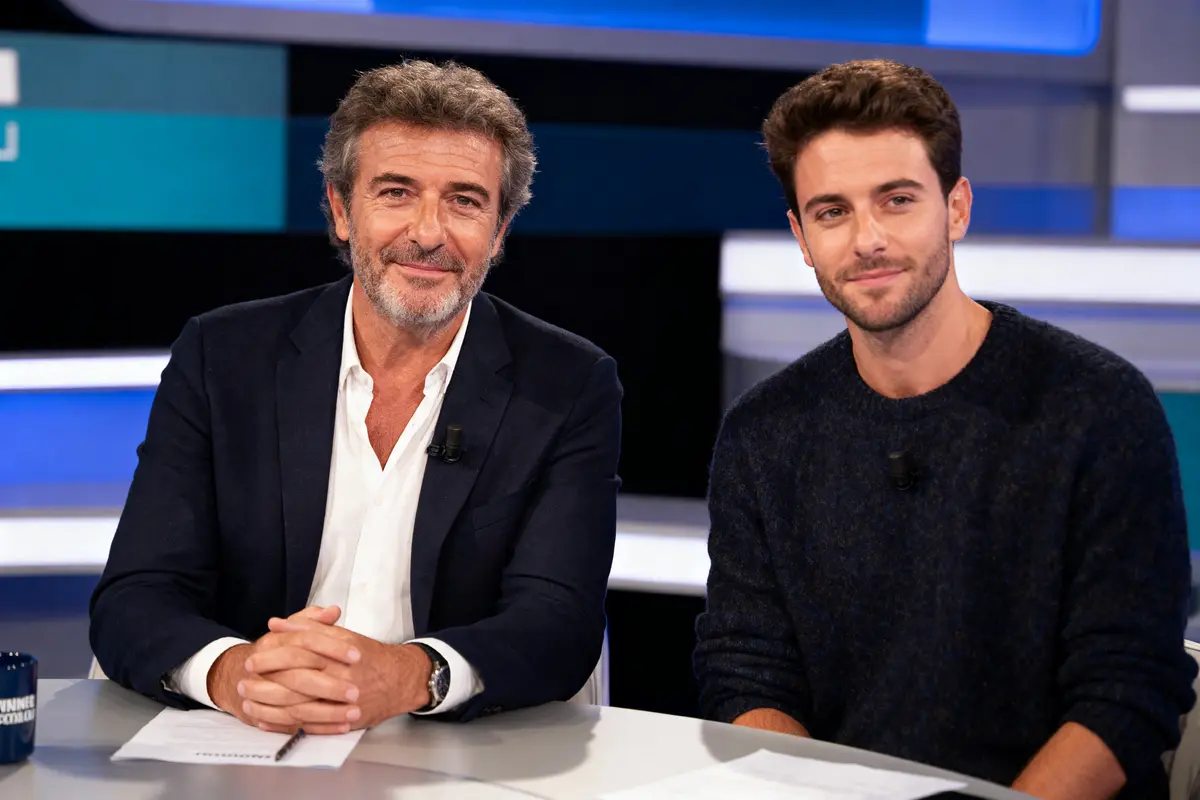 Deux hommes souriants assis à un bureau dans un studio télévisé.