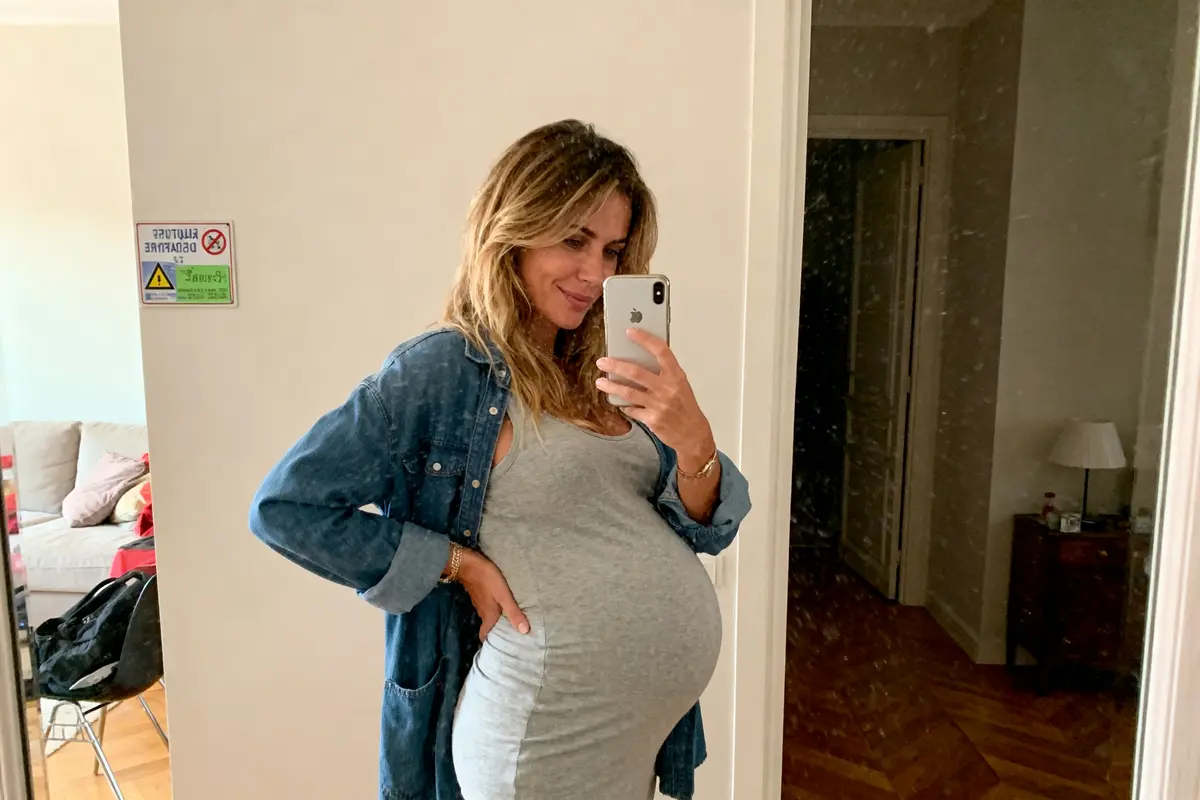 Femme enceinte souriante prenant un selfie dans un miroir, portant une robe grise et une chemise en jean.