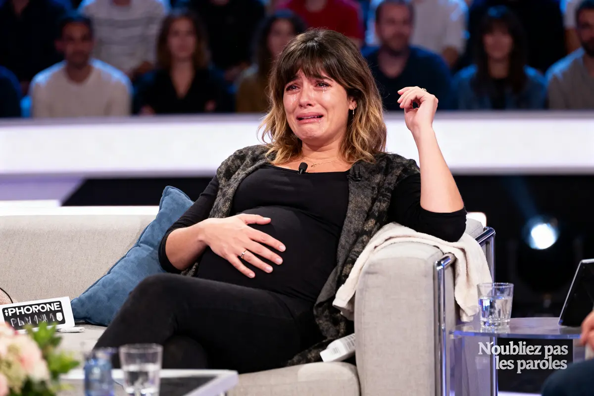 Une femme enceinte émotive assise sur un canapé lors d'une émission télévisée, spectateurs flous en arrière-plan.
