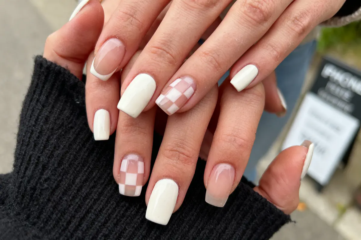 Main avec ongles manucurés, vernis blanc et motif damier, tenue par une autre main en noir.