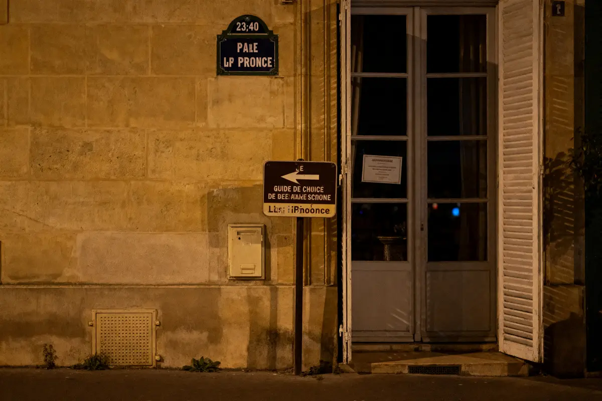 Plaque de rue bleue sur mur de pierre, panneau directionnel, entrée vitrée. Nuit, éclairage jaune.