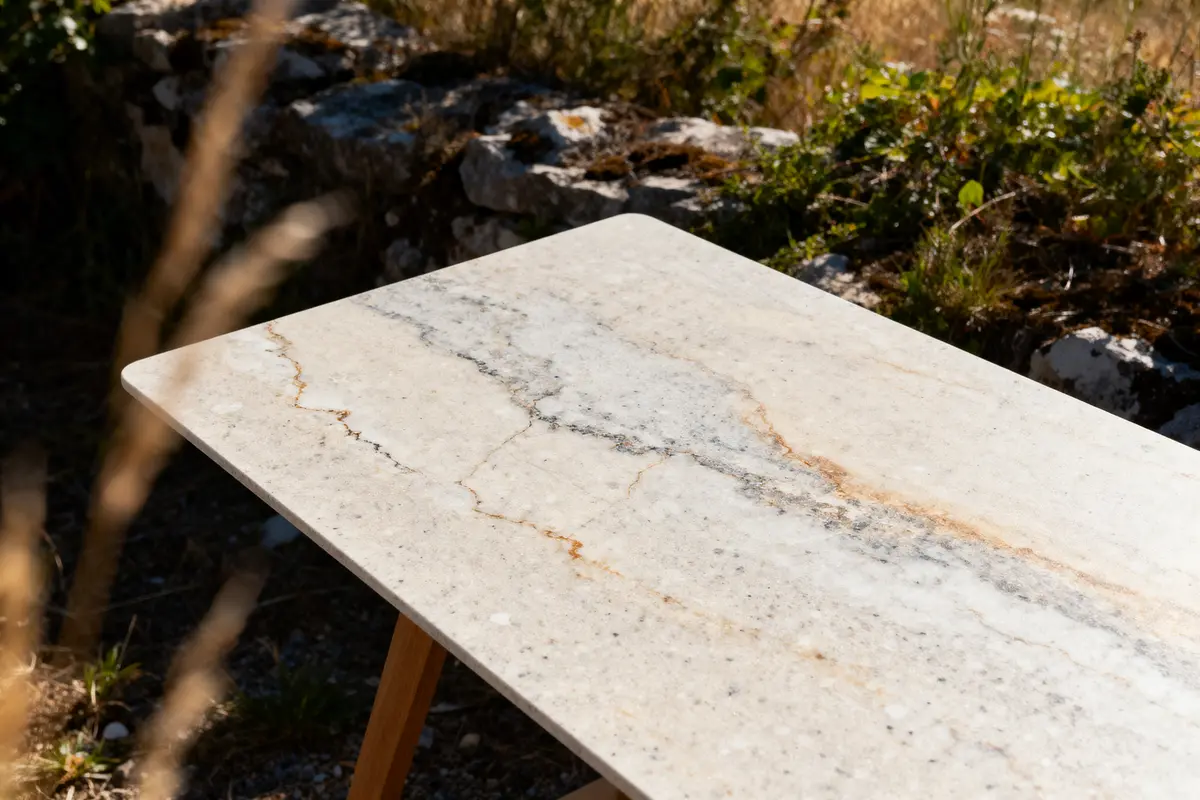 Table en marbre clair avec veines, posée à l'extérieur, ensoleillée, avec des plantes en arrière-plan.