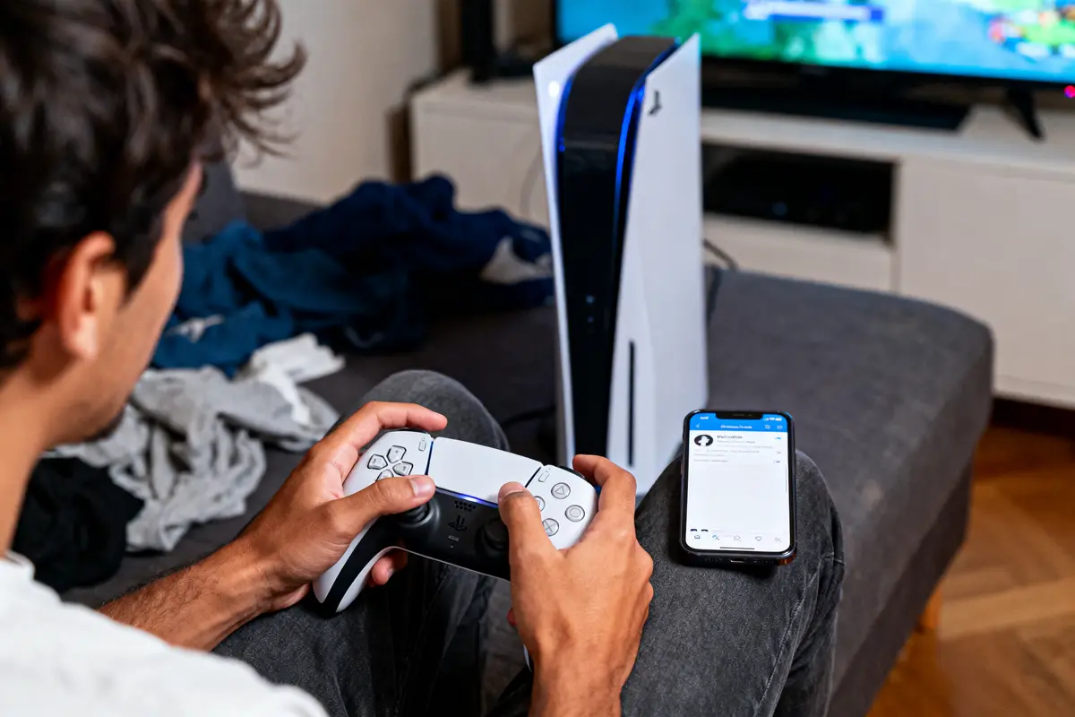 Homme jouant avec une manette de console, téléphone et télévision en arrière-plan, vêtements sur canapé.