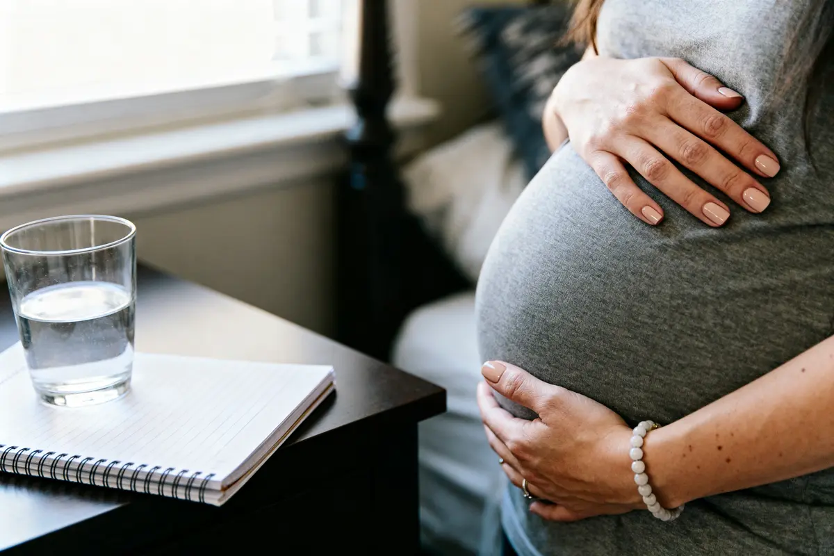 Femme enceinte en gris touchant son ventre, carnet et verre d'eau sur la table.