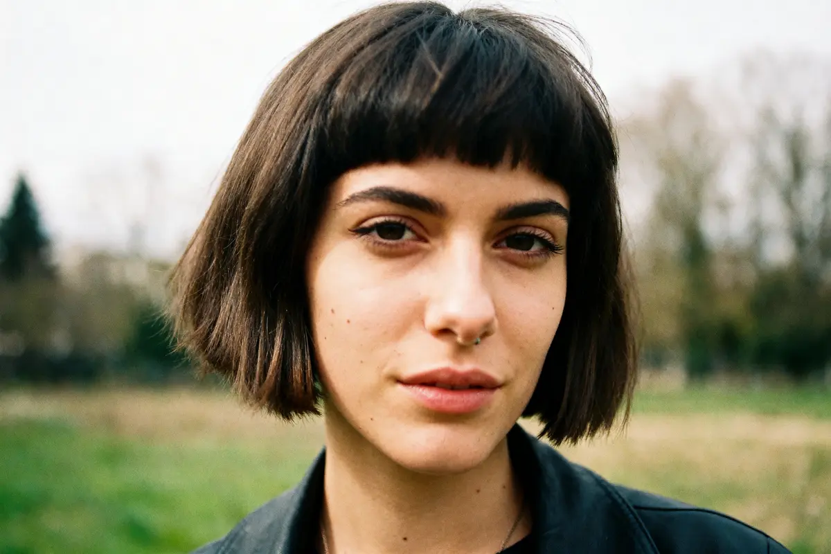 Une personne avec une coupe courte et un septum, posant en extérieur flou en arrière-plan.
