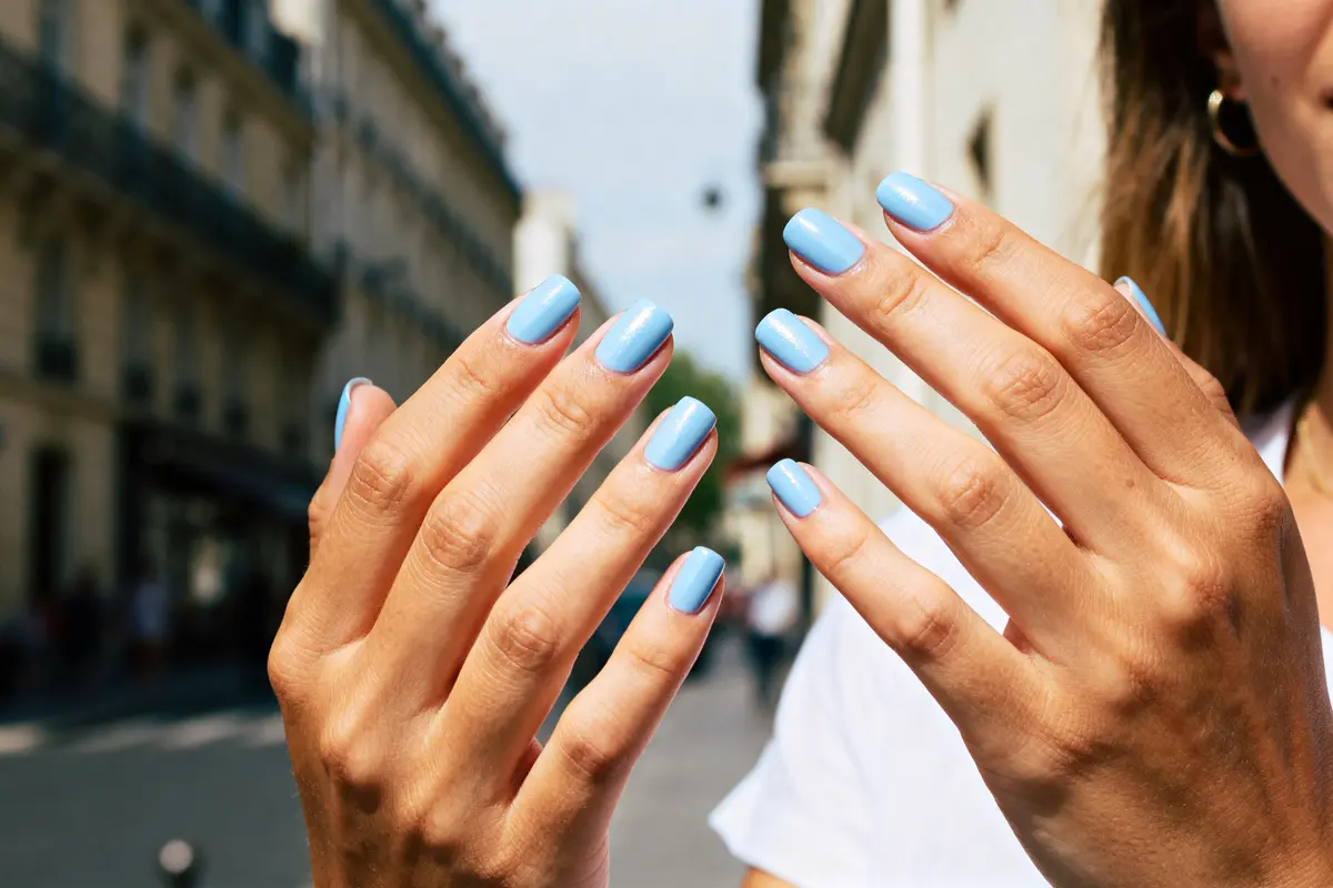 Mains avec ongles bleus pastel dans une rue parisienne ensoleillée